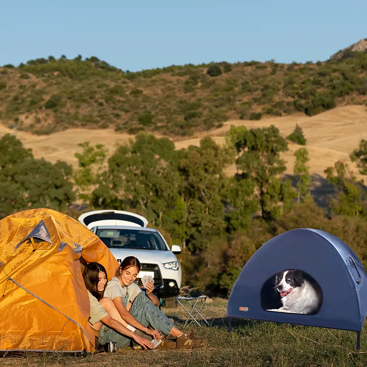 Two people smile while tying their shoes beside an orange tent in a grassy area. Nearby, a dog is comfortably inside a small blue Dog Tent with Sun Protection. A car is parked behind them, and hills dotted with trees are visible in the background.