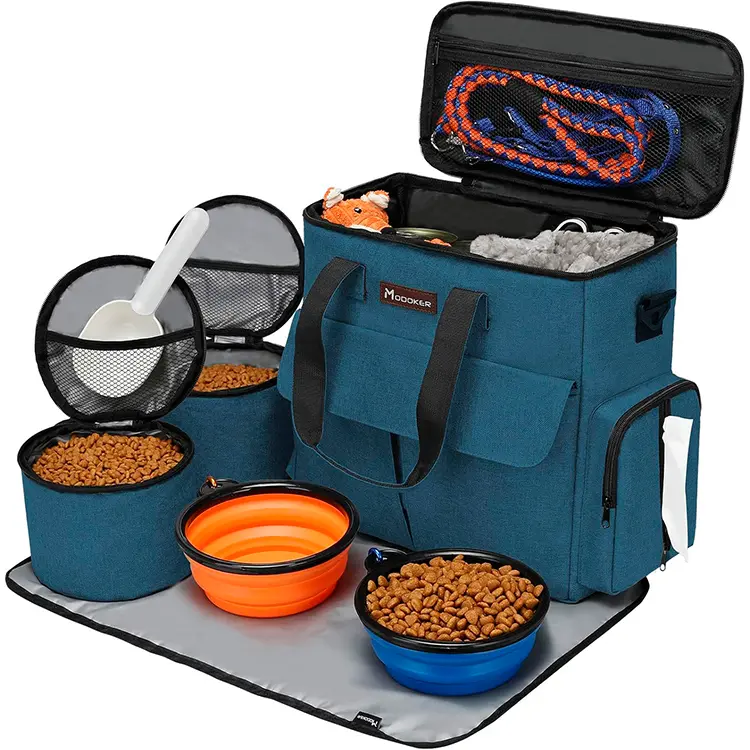 The Dog Travel Bag is open, revealing storage compartments with pet essentials: collapsible food bowls with kibble, portable containers, a white scoop, and leashes. A gray mat is laid out in front.