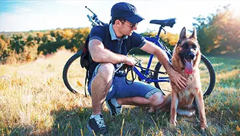 A man wearing glasses and a cap kneels beside a German Shepherd. They are outdoors in a grassy field, with a bicycle in the background. The man is smiling and petting the dog, and the sun is shining brightly.