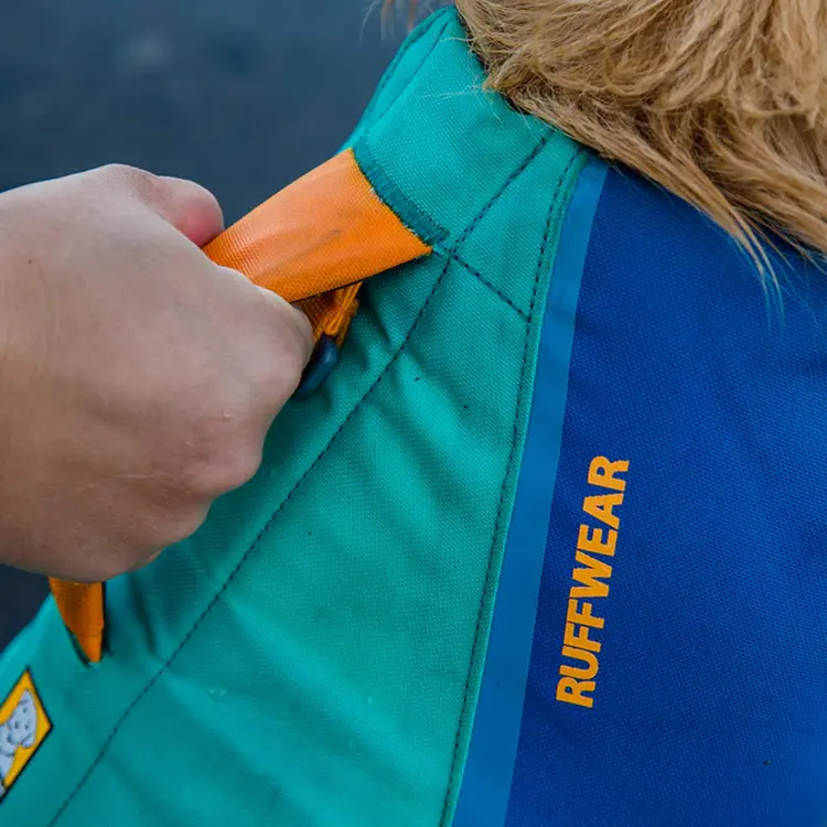 A close-up shows a persons hand holding the handle of a blue and green Dog Life Jacket worn by a dog, with the orange text RUFFWEAR visible on it.