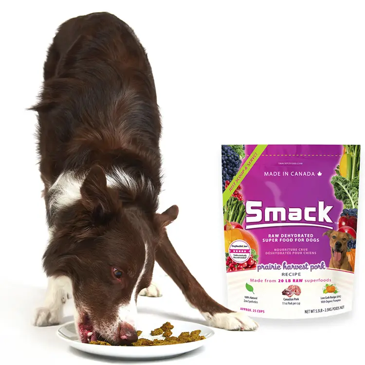 A brown and white dog eats from a bowl on the floor beside a bag of Smack Pet Food, labeled prairie harvest pork recipe, with images of vegetables and a small dog.