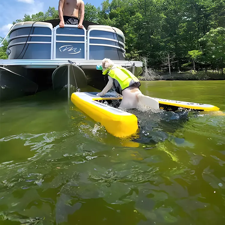 A dog wearing a yellow life jacket climbs onto a Dog Water Ramp attached to a yellow inflatable raft in the water near a pontoon boat. A person sits on the boats edge, with green trees and a blue sky in the background.
