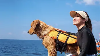 A woman in a visor smiles while holding a wet Golden Retriever wearing an orange life jacket. They are on a boat near a vast blue ocean under a bright sky.