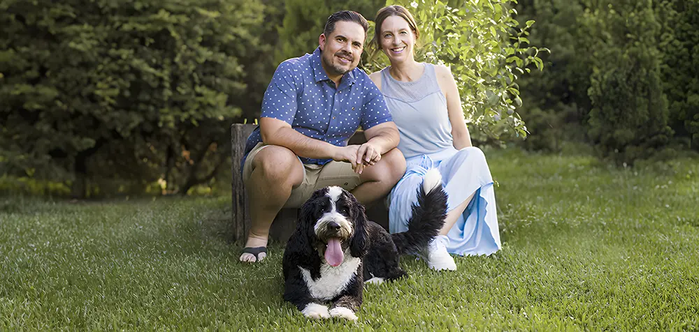 A man and a woman sit on a wooden bench in a lush garden, smiling. A large, fluffy black and white dog lies happily on the grass in front of them. The woman wears a light blue dress, and the man wears a patterned blue shirt.