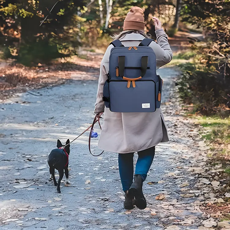A person in a light coat and beanie walks a small dog on a leash along a wooded, leaf-covered path. They carry their Backpack for Dog Accessories in blue and wave with one hand, surrounded by trees.