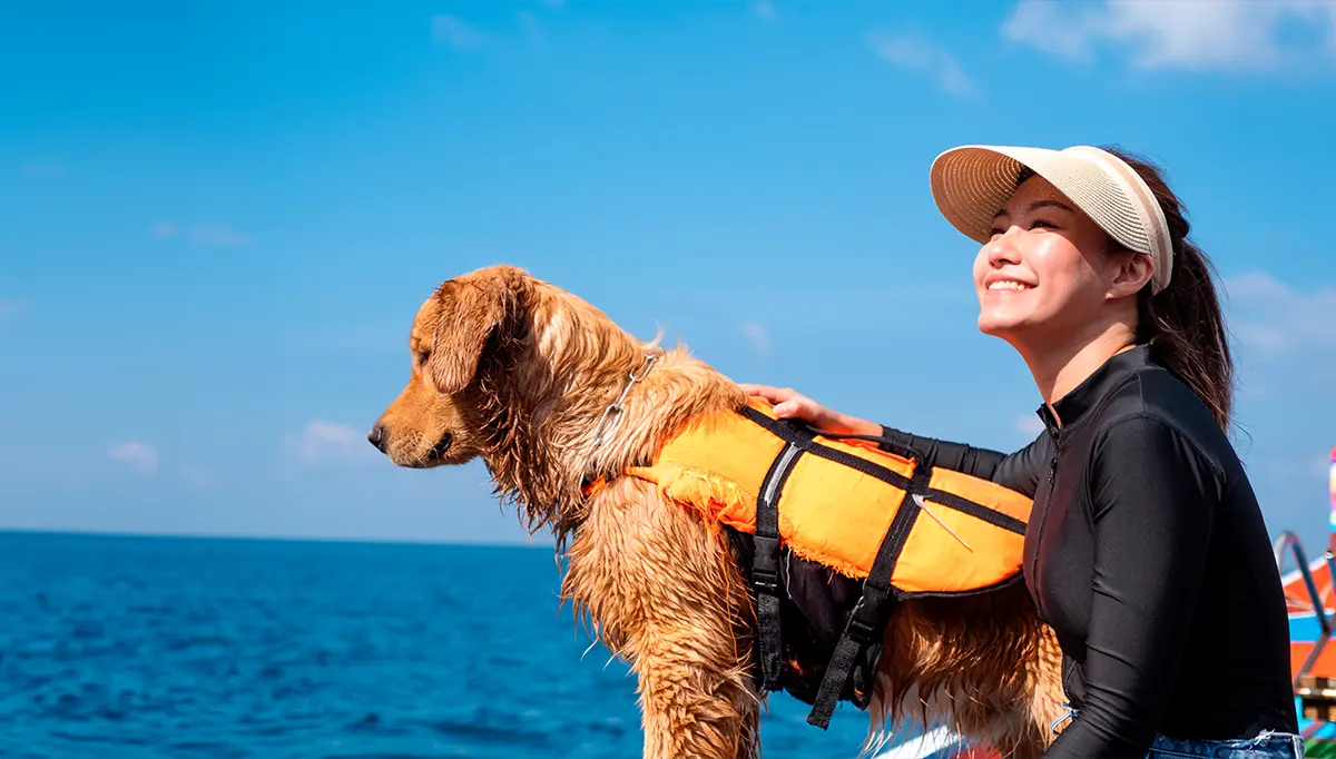 A woman in a visor smiles while holding a wet Golden Retriever wearing an orange life jacket. They are on a boat near a vast blue ocean under a bright sky.