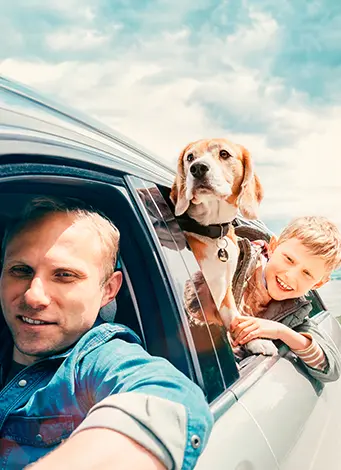 A man driving a car takes a selfie with a beagle and a smiling boy. The beagle is leaning out of the car window, and both appear happy under a partly cloudy sky. The scene suggests a cheerful road trip.