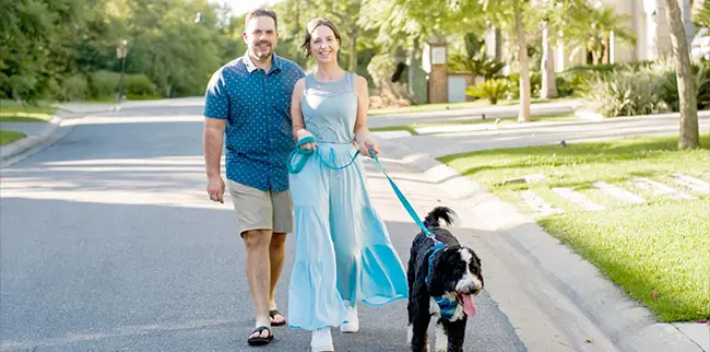 A couple walks down a suburban street with a black and white dog on a leash. The man wears a blue shirt and shorts, and the woman wears a light blue dress. The street is lined with grass and trees under a clear sky.