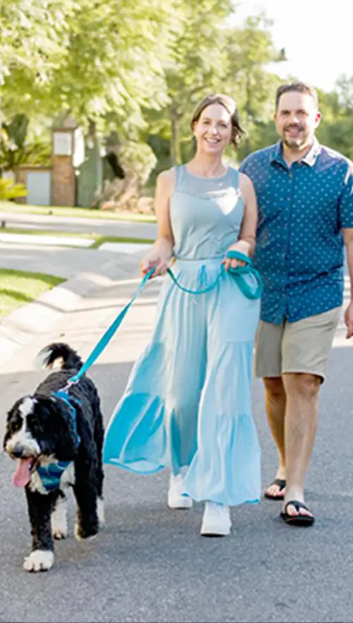 A couple walks down a suburban street with a black and white dog on a leash. The man wears a blue shirt and shorts, and the woman wears a light blue dress. The street is lined with grass and trees under a clear sky.