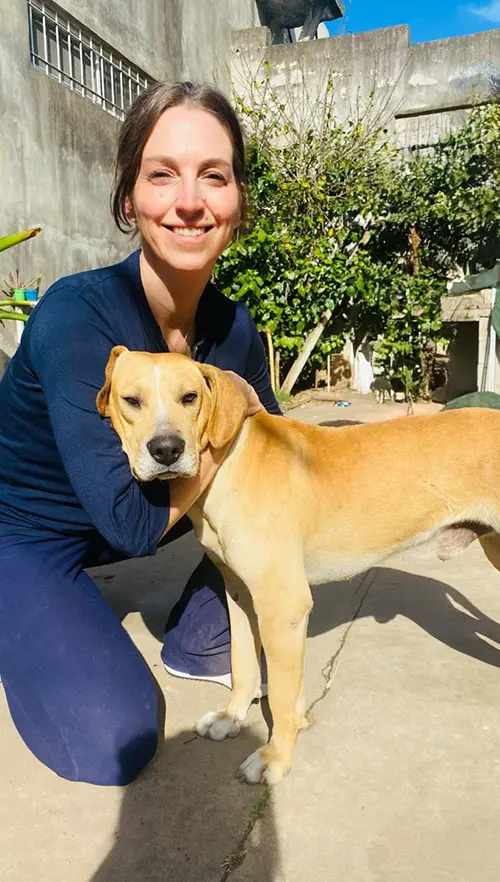 A woman with short dark hair is smiling and hugging a light brown and white dog outside. They are standing on a concrete path with greenery and a building in the background. The sun is shining brightly.
