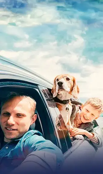 A man driving a car takes a selfie with a beagle and a smiling boy. The beagle is leaning out of the car window, and both appear happy under a partly cloudy sky. The scene suggests a cheerful road trip.