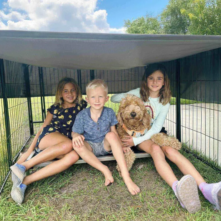 Three children and a fluffy brown dog sitting comfortably inside a large portable play area with a shaded canopy, set up outdoors on grass. The sturdy, versatile playpen creates a safe and relaxing space for pets and kids